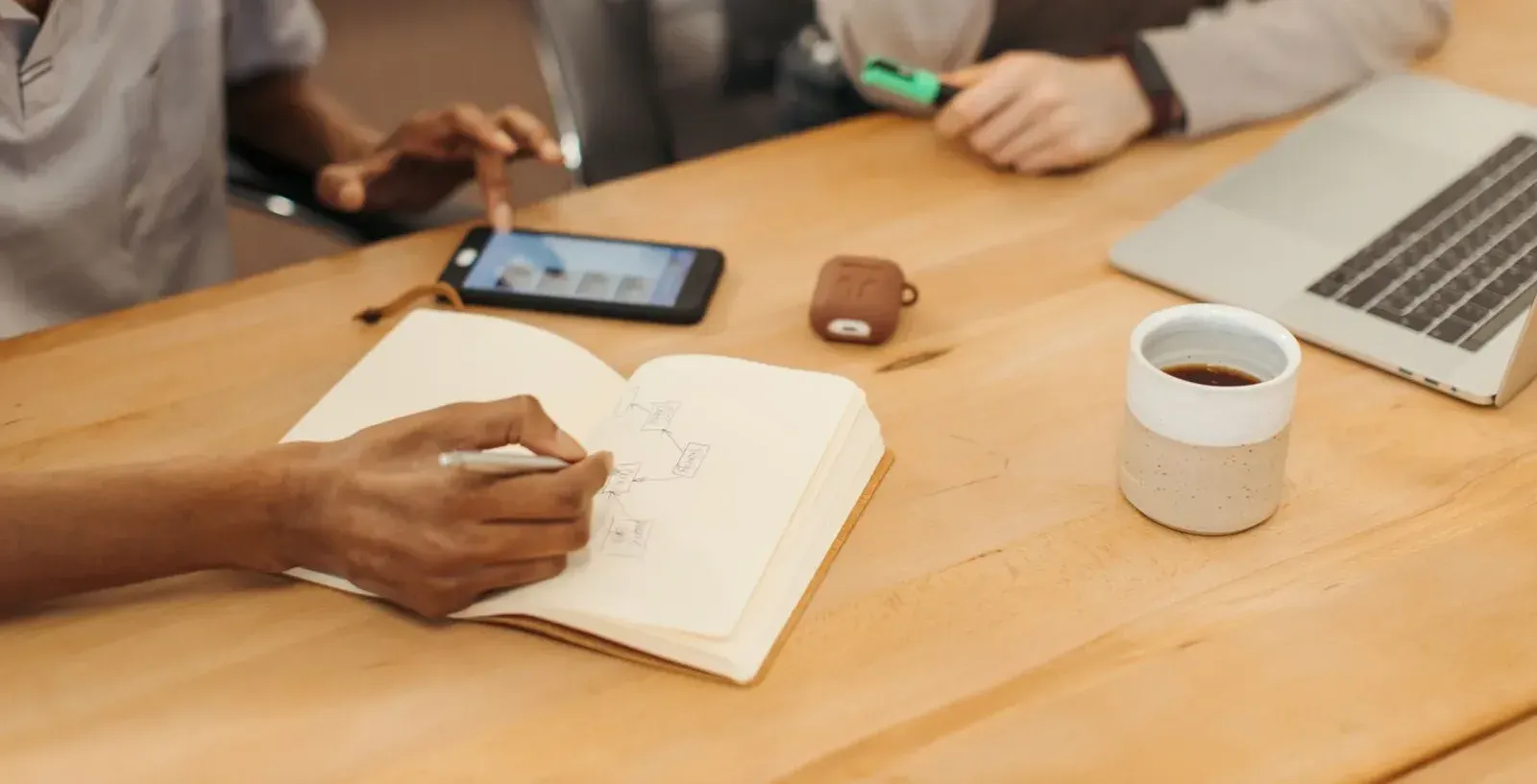 Mão desenhando o fluxo de um processo em um caderno, sobre uma mesa com celular, caneca de café e notebook